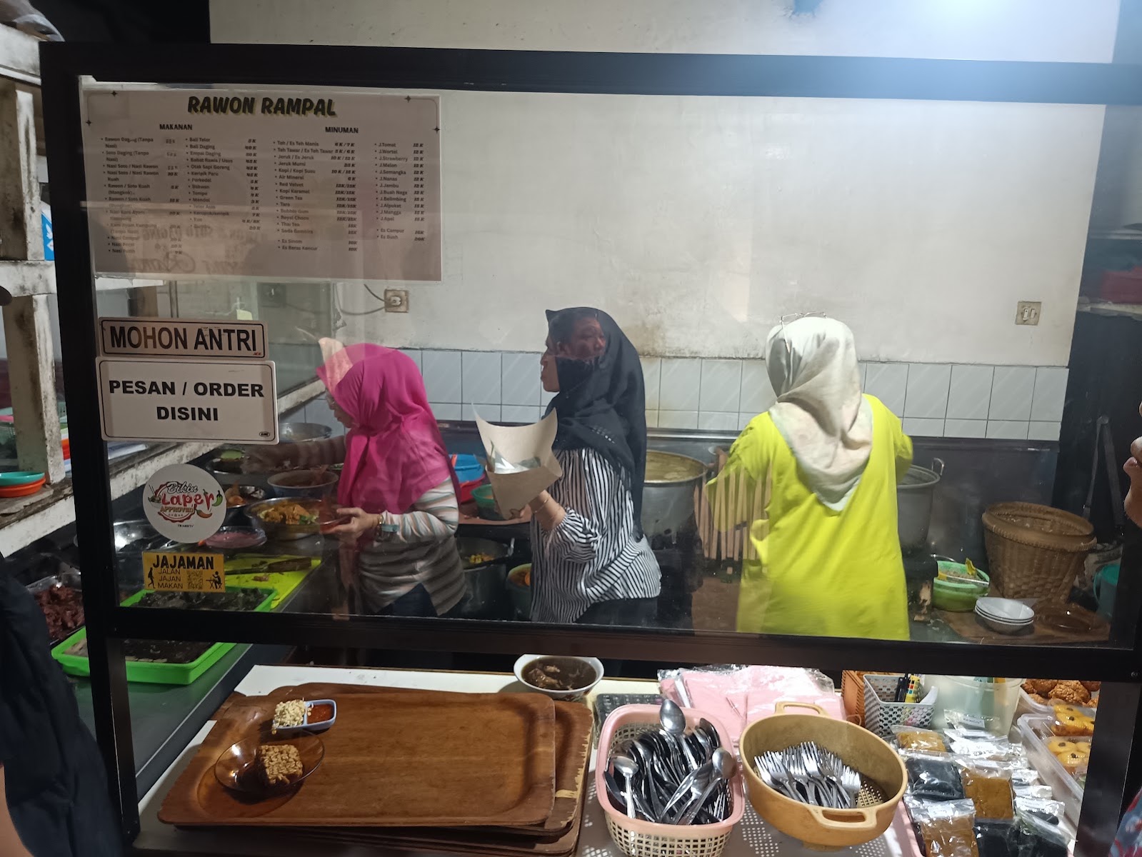 Konter pesanan Rawon Rampal dengan papan bertuliskan mohon antri dan pesan order di sini, tiga petugas berkerudung menyiapkan pesanan di baliknya.
