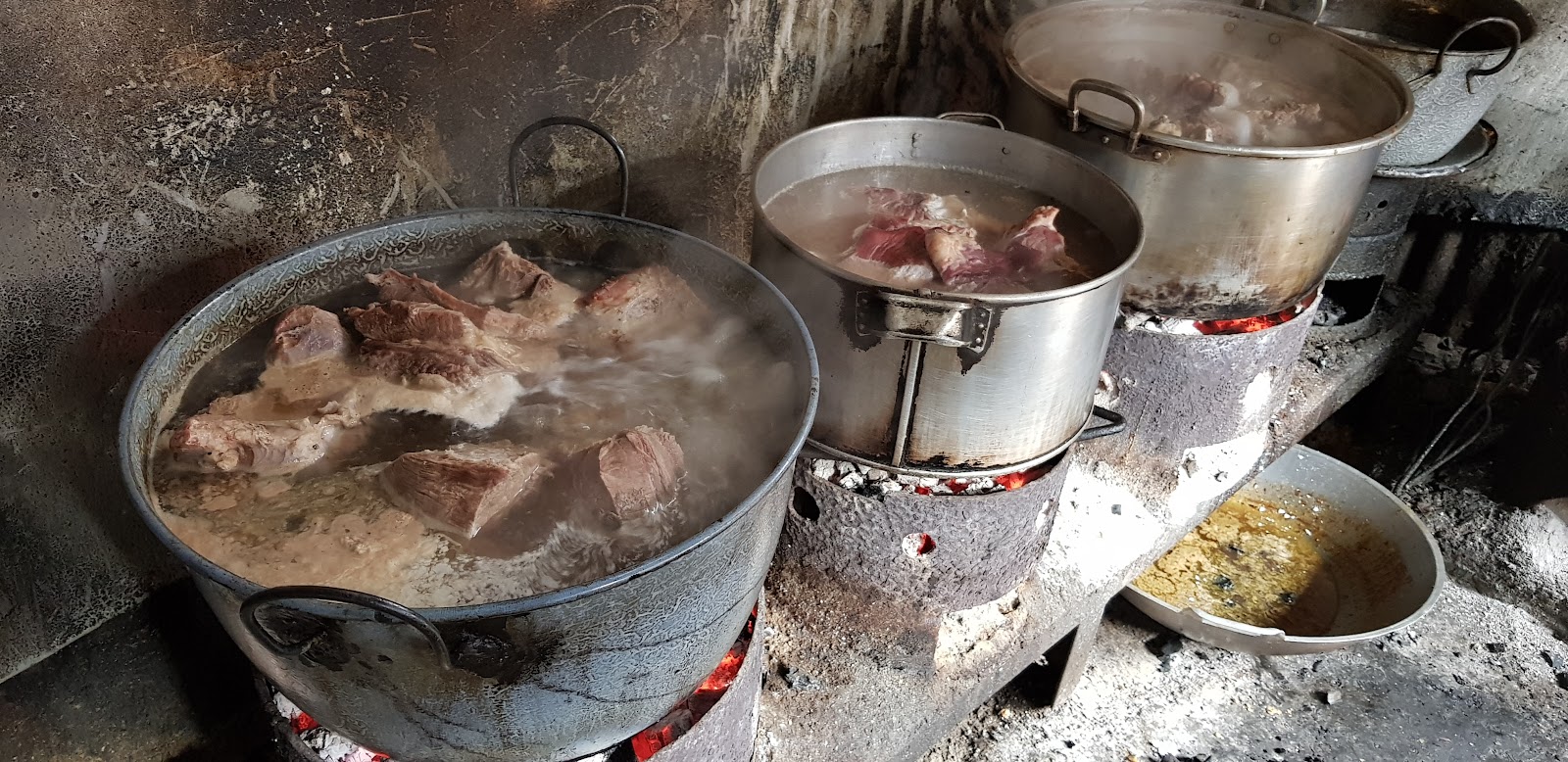 Dapur warung: dua panci logam besar berisi kuah mendidih di atas tungku, dengan potongan daging di dalamnya.