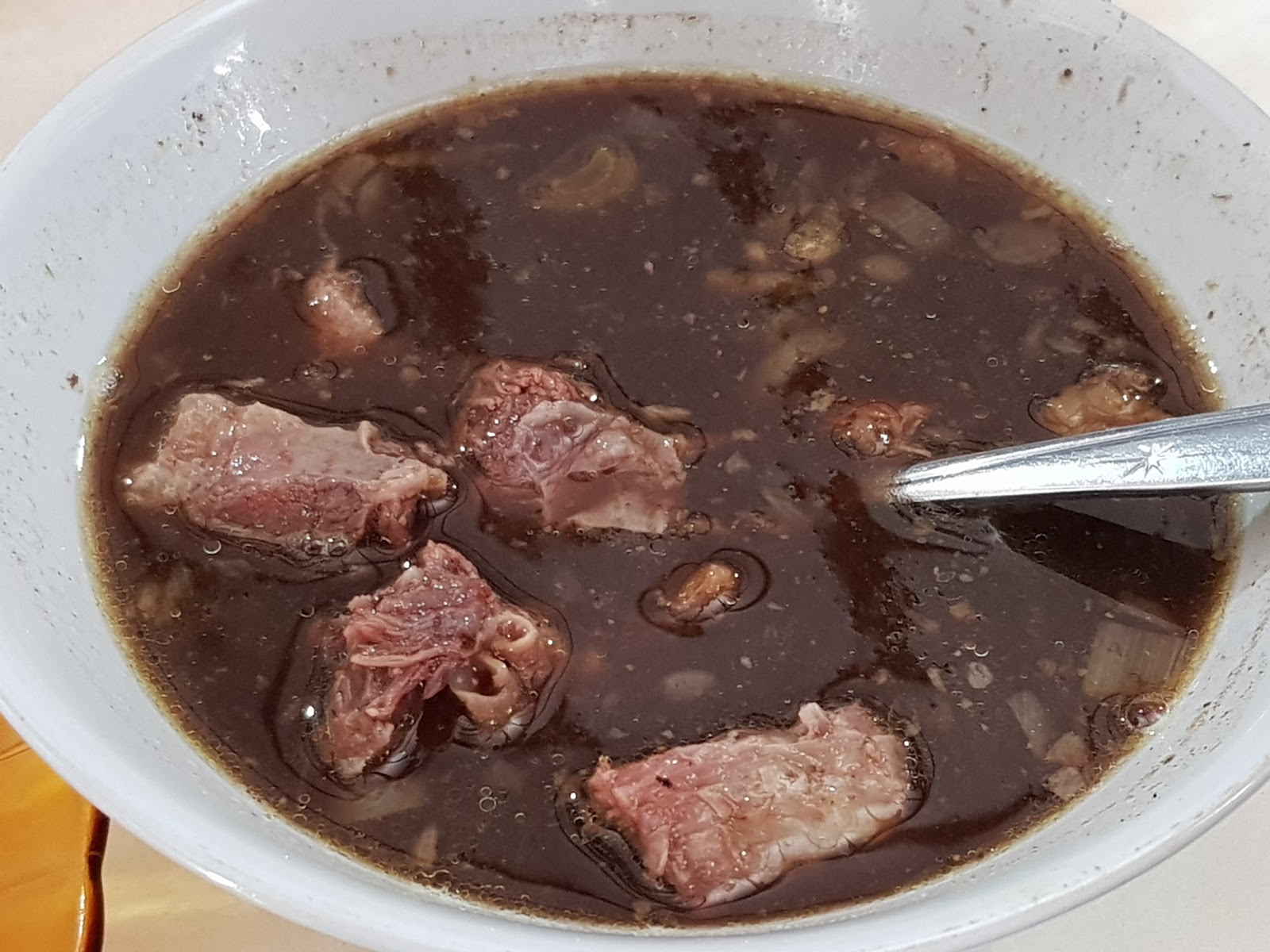 Semangkuk rawon berkuah hitam pekat dengan potongan daging sapi besar dan sendok logam bersandar di tepi mangkuk.